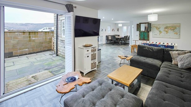 Un salón moderno con un sofá gris, una mesa de centro de madera y un televisor. Hay una zona de comedor con una mesa y sillas, y un amplio ventanal con vistas a un muro de ladrillo.