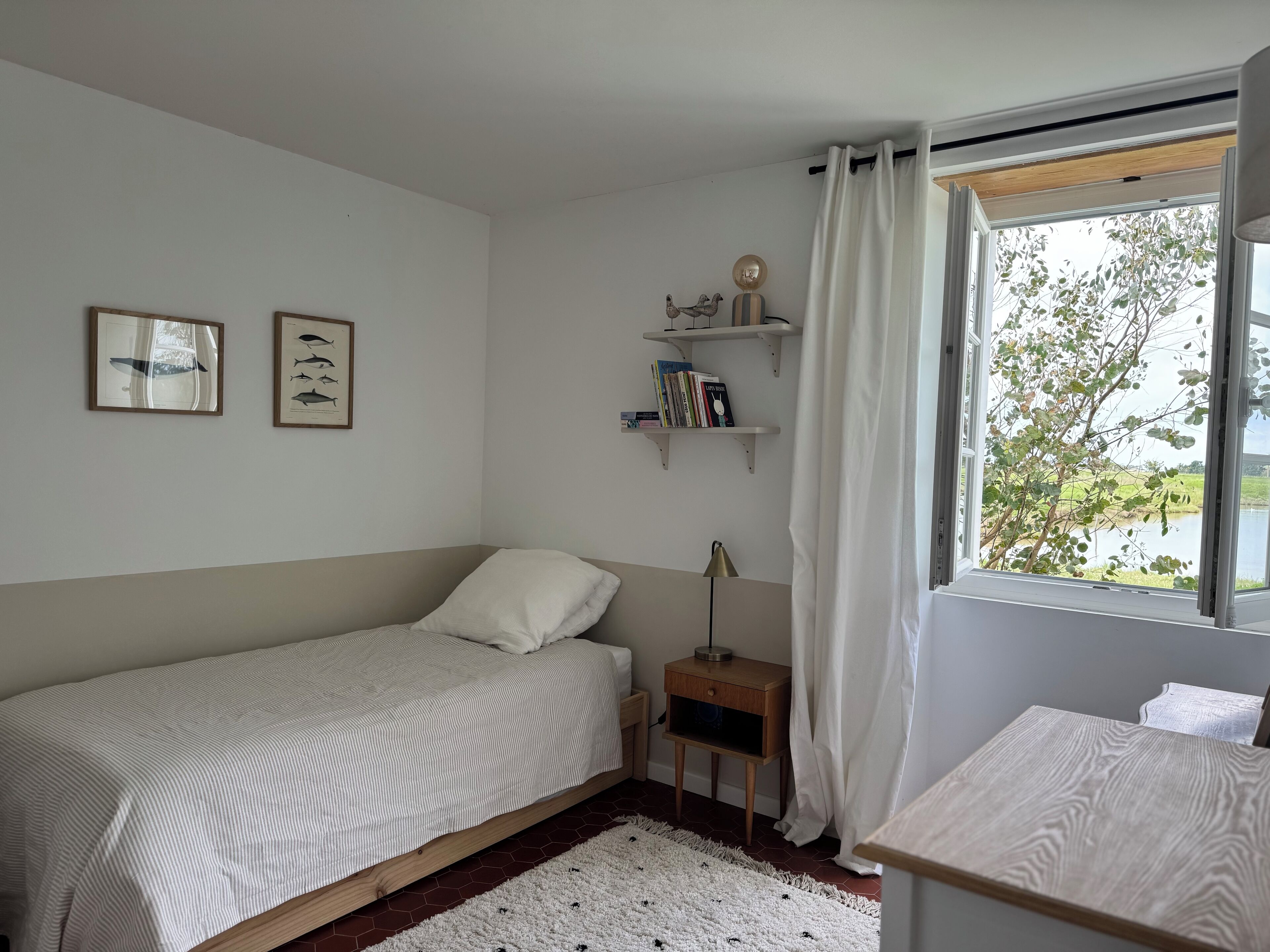 A bedroom with a single bed, a nightstand, a lamp, a window with a view of greenery, and two framed fish illustrations on the wall.