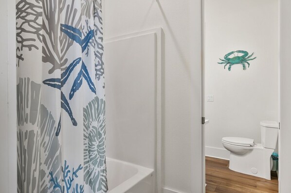 A bathroom with a white bathtub, a toilet, and a shower curtain with a leaf pattern. - Tidewater 4 Bedroom Home by RedAwning (Port Bolivar)
