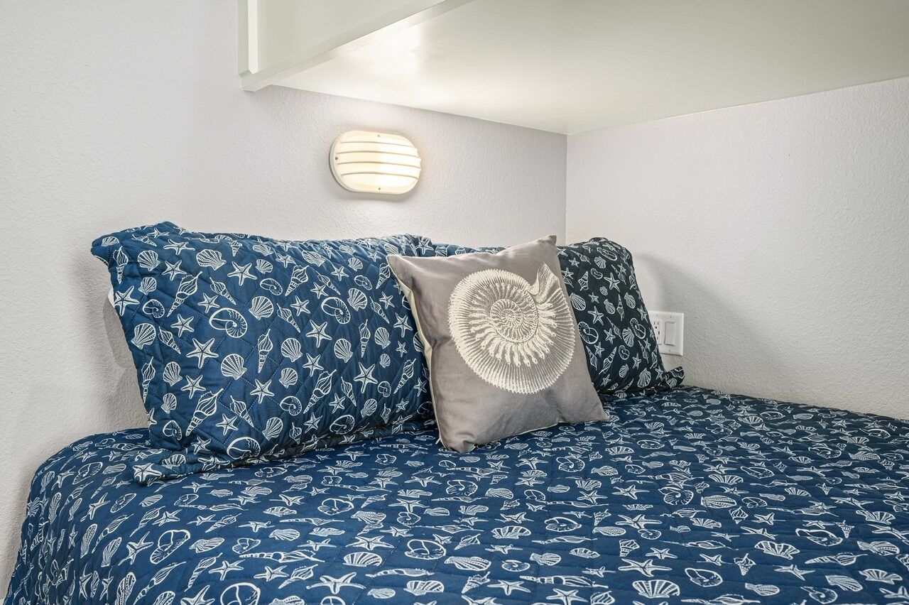 Una cama con colcha azul y blanca de temática náutica, dos almohadas con un diseño parecido y una lámpara de pared.