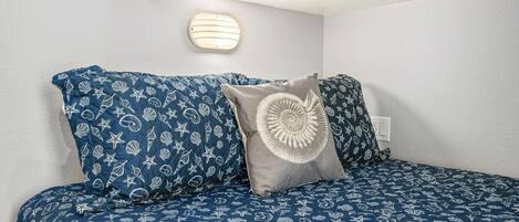 Una cama con colcha azul y blanca de temática náutica, dos almohadas con un diseño parecido y una lámpara de pared.