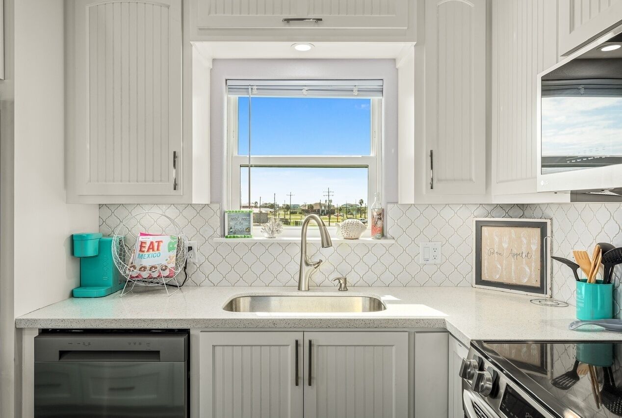A kitchen with white cabinets, a stainless steel sink, a window with a view, and a teal coffee maker.