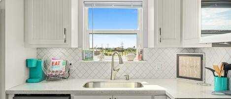 A kitchen with white cabinets, a stainless steel sink, a window with a view, and a teal coffee maker.