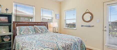 A bedroom with a bed, a ceiling fan, a window with blinds, a door with a glass panel, and a wall-mounted mirror.