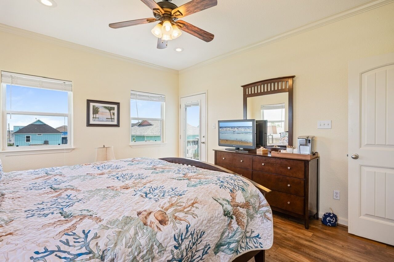 Ein Schlafzimmer mit Bett, Deckenventilator, Kommode, Fernseher und Fenstern mit Blick auf den Strand.