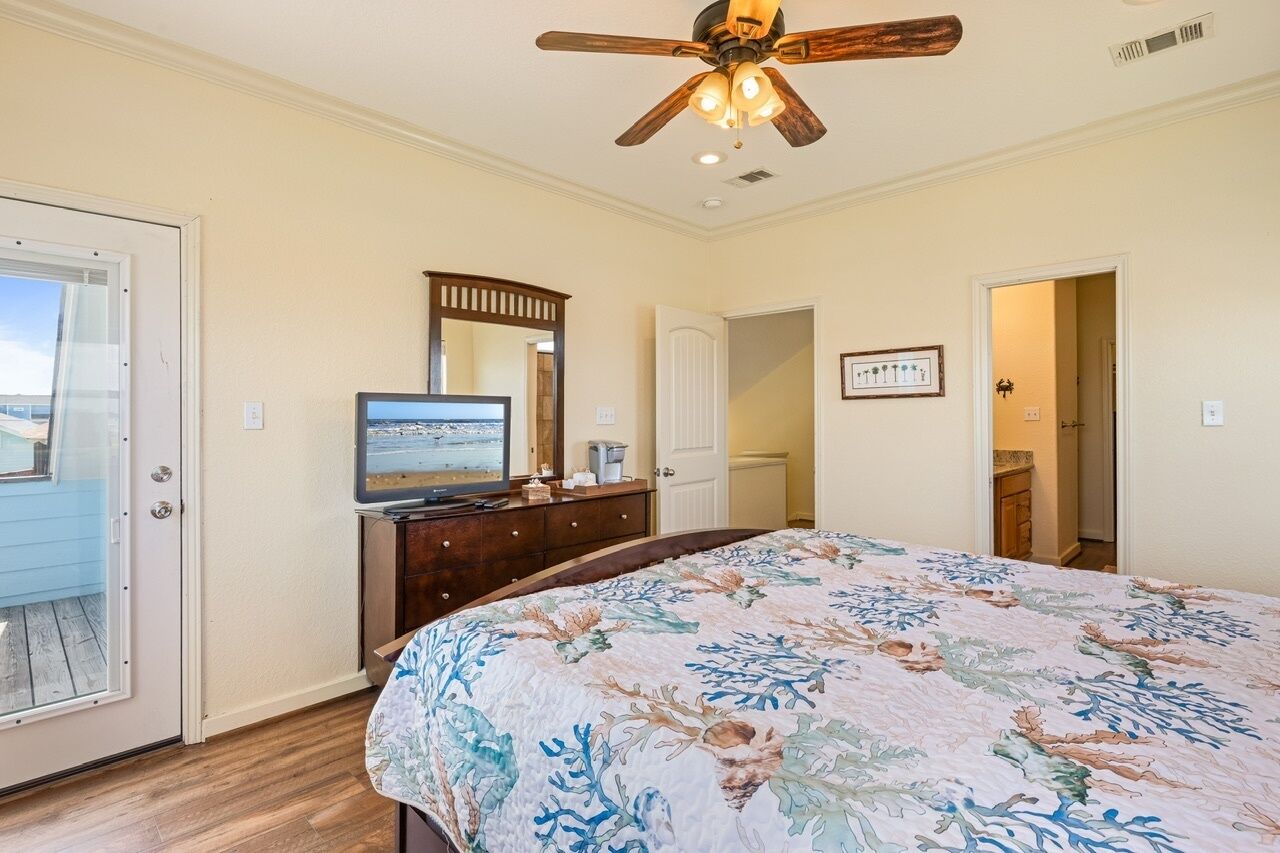 A bedroom with a bed, a ceiling fan, a dresser, a television, and a door leading to another room.