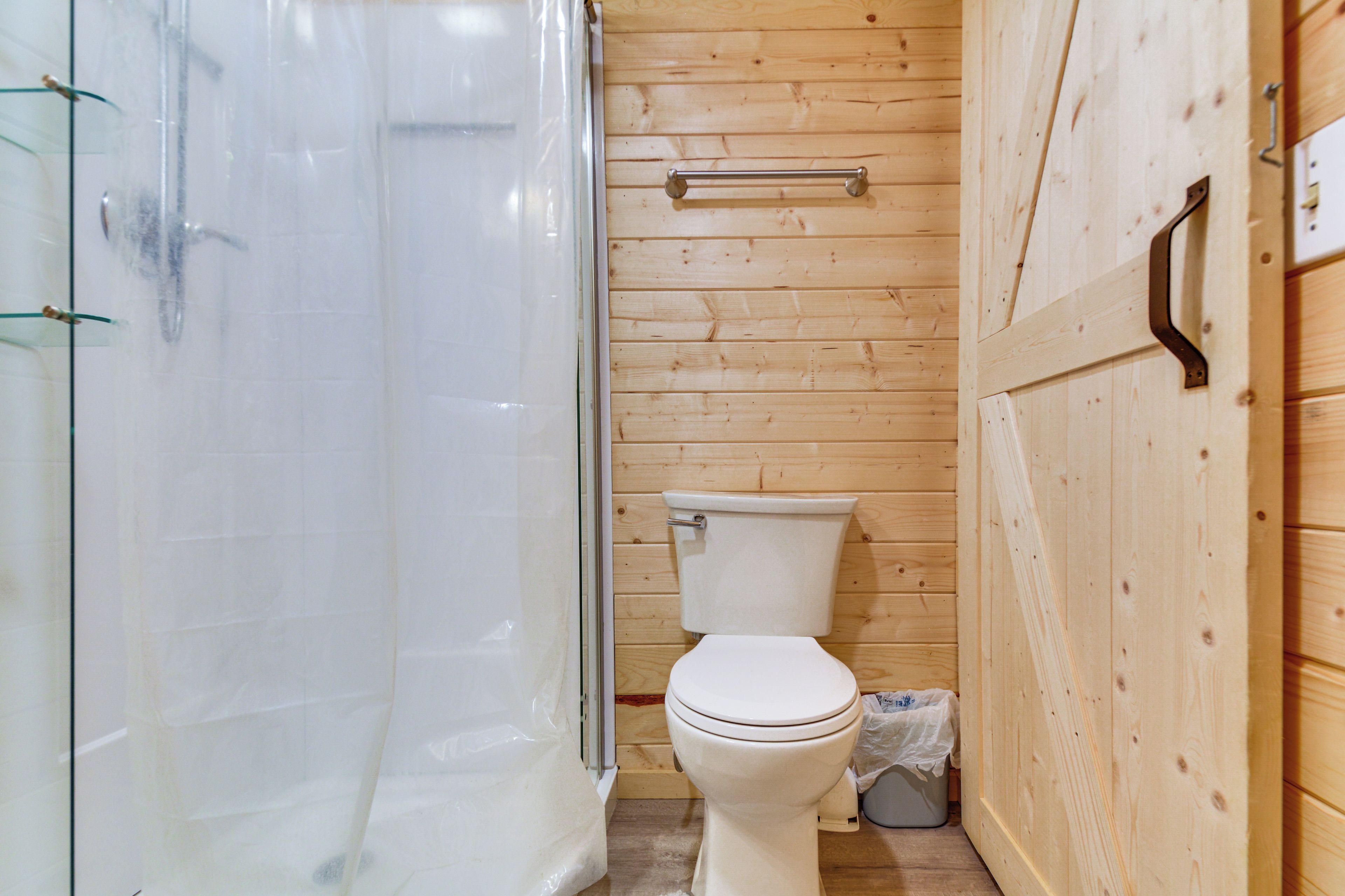 A wooden-paneled bathroom with a glass shower enclosure, a toilet, and a small trash bin.
