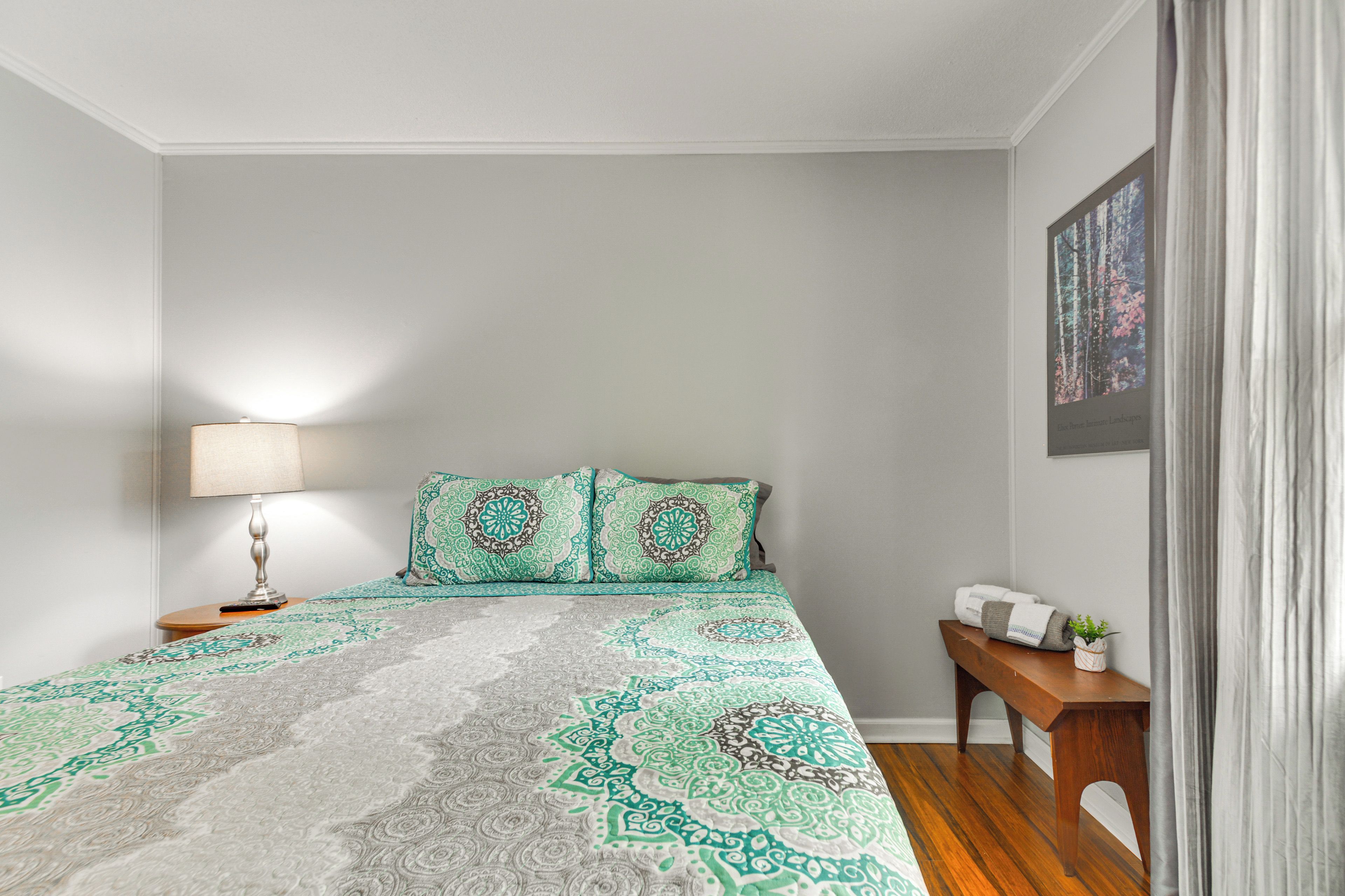 Un dormitorio con una cama, una mesita de noche con lámpara, un banco y un cuadro enmarcado en la pared.