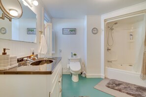 Une salle de bain avec un lavabo, des toilettes et une baignoire. Il y a des horloges au mur, une serviette accrochée à la porte et une petite plante posée sur les toilettes.
