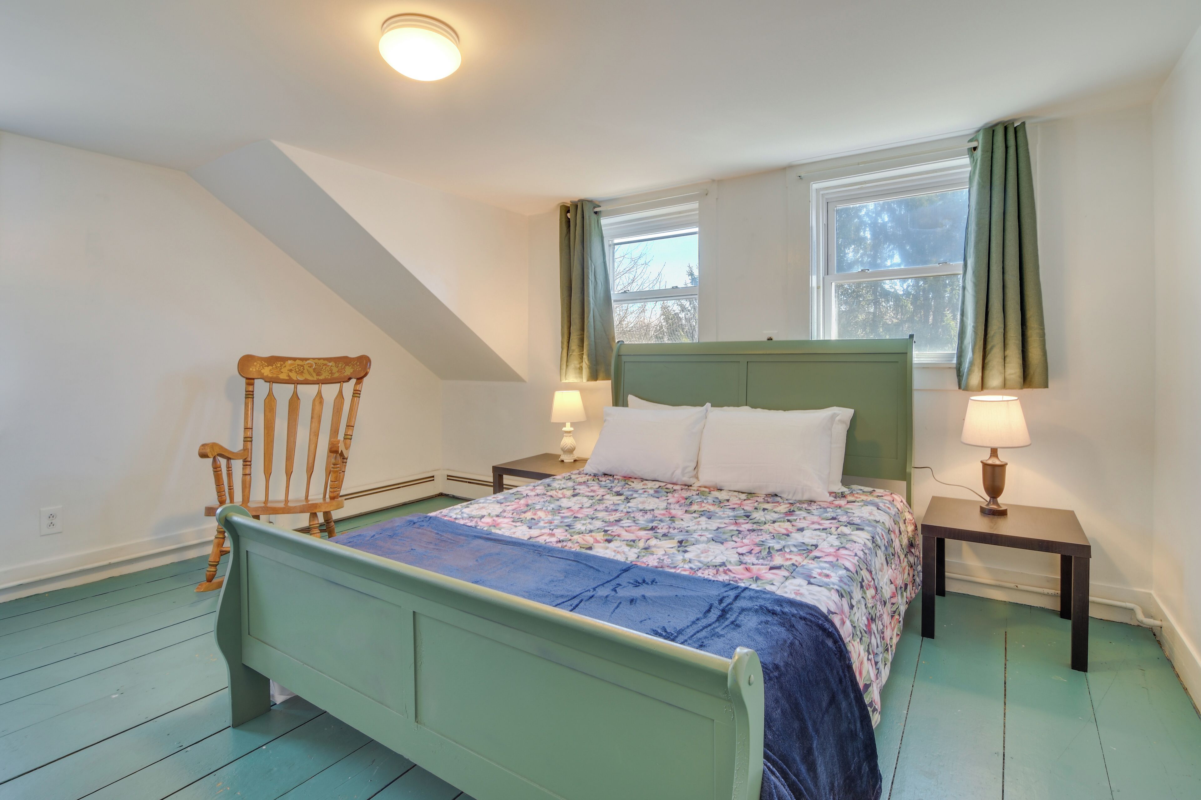 A bedroom with a green bed, a wooden chair, two bedside tables with lamps, and a window with curtains.