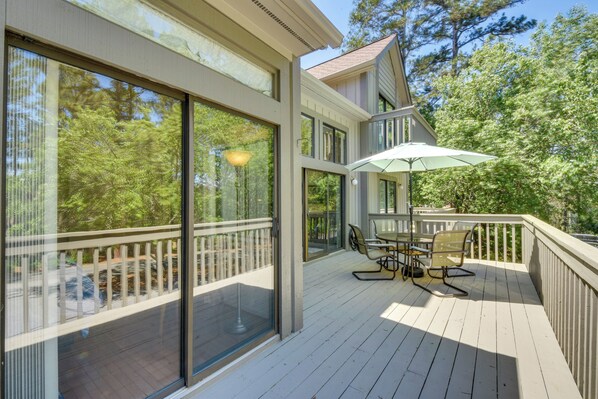 A wooden deck with a table and chairs, large windows, and a view of trees.