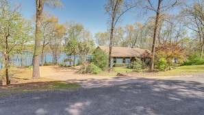 A single-story house with a gravel driveway and a lawn, surrounded by trees and a body of water. - Tranquil Lakefront Greers Ferry Escape w/ Fire Pit (Higden)