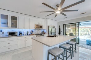 Una cocina moderna con gabinetes blancos, una isla central con taburetes altos, un ventilador de techo y un ventanal con vistas al exterior.