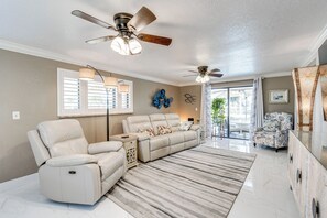 Amplia sala de estar con un sofá color beige, un sillón reclinable y un tapete estampado. Hay ventiladores de techo, un ventanal con persianas y una puerta que da a otra habitación.