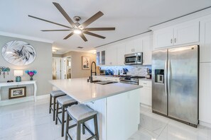 Una cocina moderna con armarios blancos, una isla grande con taburetes de bar, electrodomésticos de acero inoxidable y un ventilador de techo.