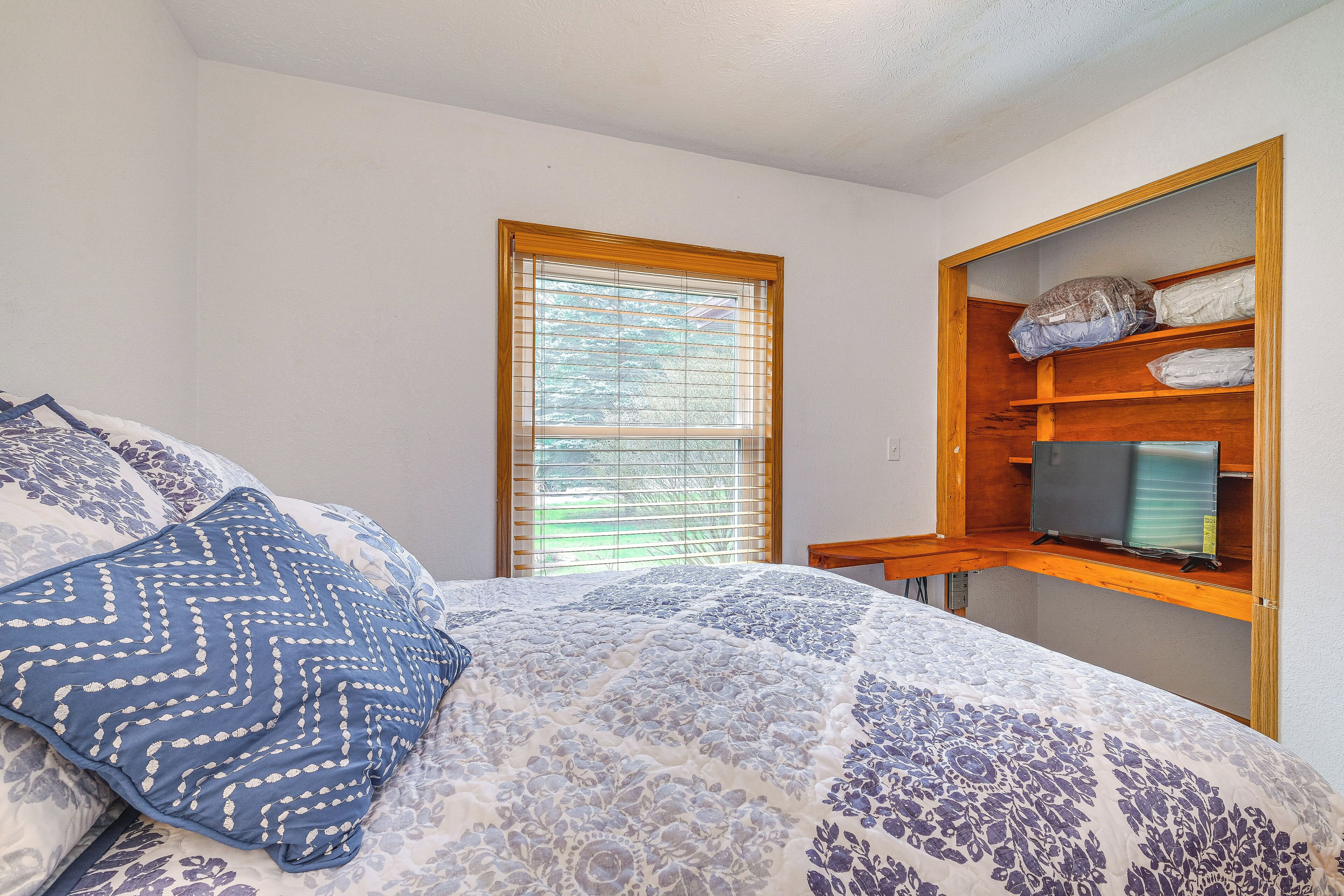 Un dormitorio con cama, ventana con persianas, estante de madera con objetos y televisor.