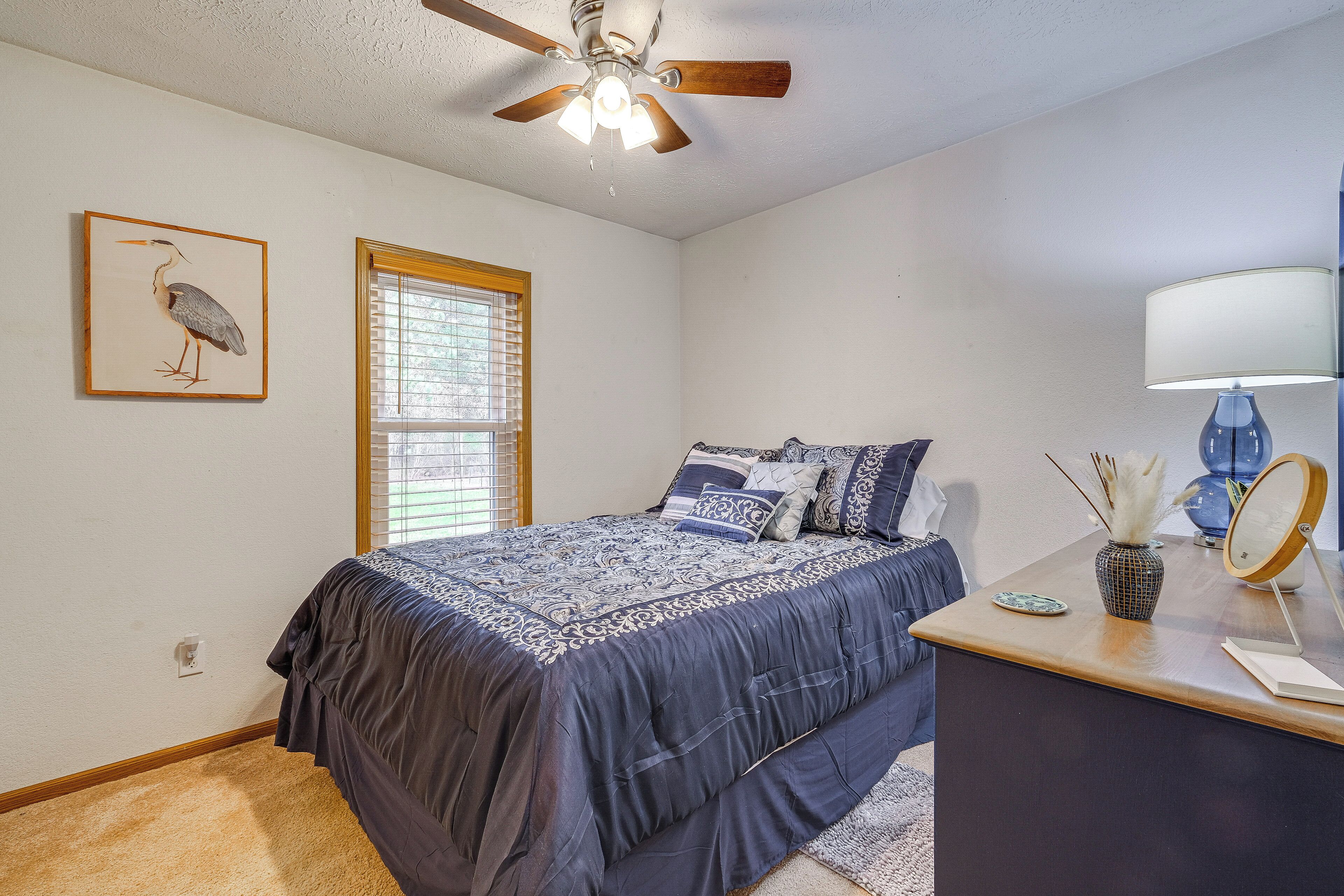 Une chambre à coucher avec un lit, un ventilateur de plafond, une fenêtre avec des stores, une table de chevet avec une lampe et des tableaux encadrés accrochés au mur.