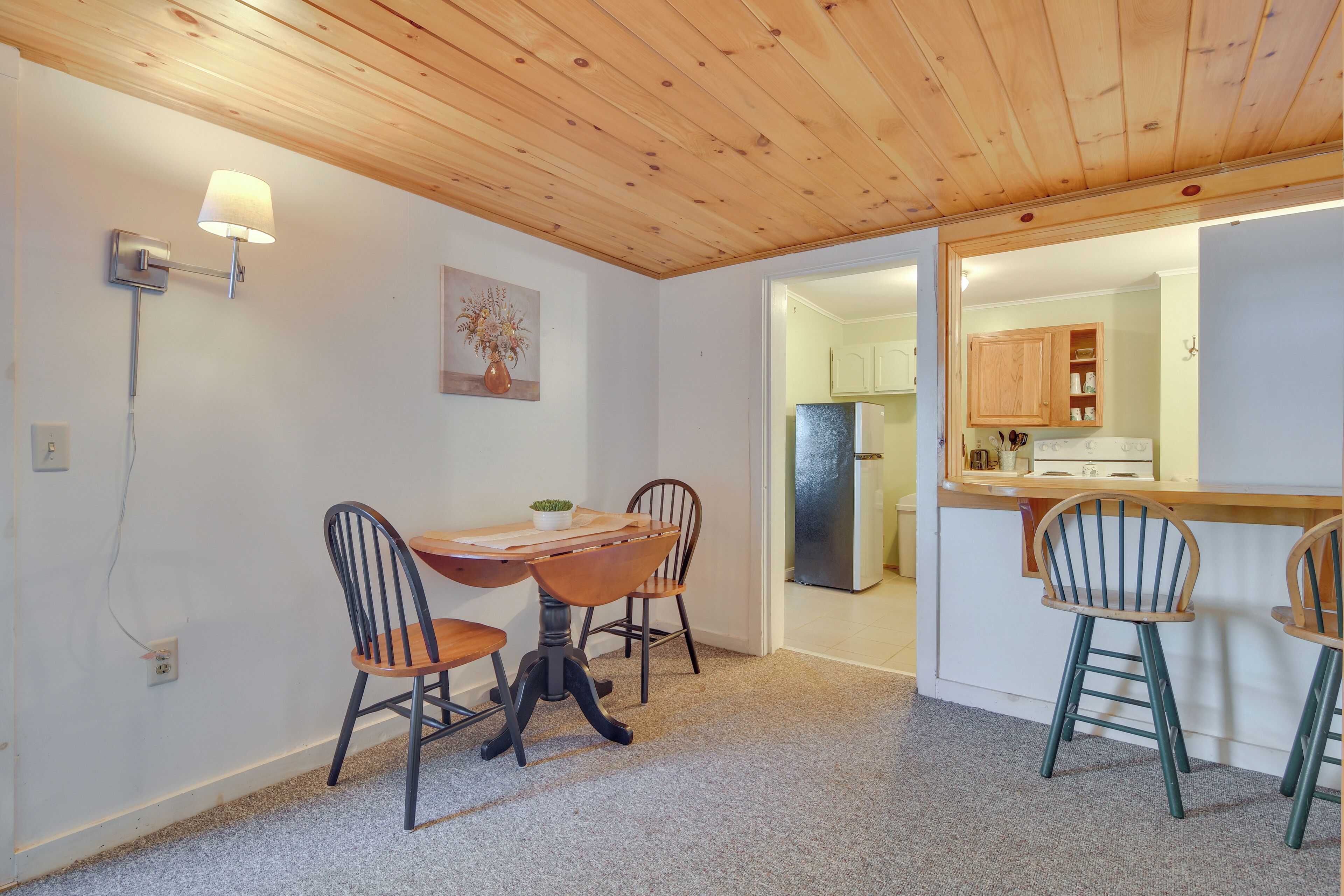 A dining area with a wooden ceiling, a table with chairs, and a kitchen visible through an open doorway.