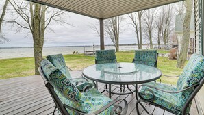 A covered outdoor seating area with a glass table and patterned chairs, overlooking a grassy area and a body of water. - Family-friendly Houghton Lake House: Steps to Lake (Houghton Lake)