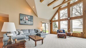 A spacious living room with a large sectional sofa, a wooden coffee table, and a colorful ottoman. A large window offers a view of the outdoors. - Family-friendly Houghton Lake House: Steps to Lake (Houghton Lake)