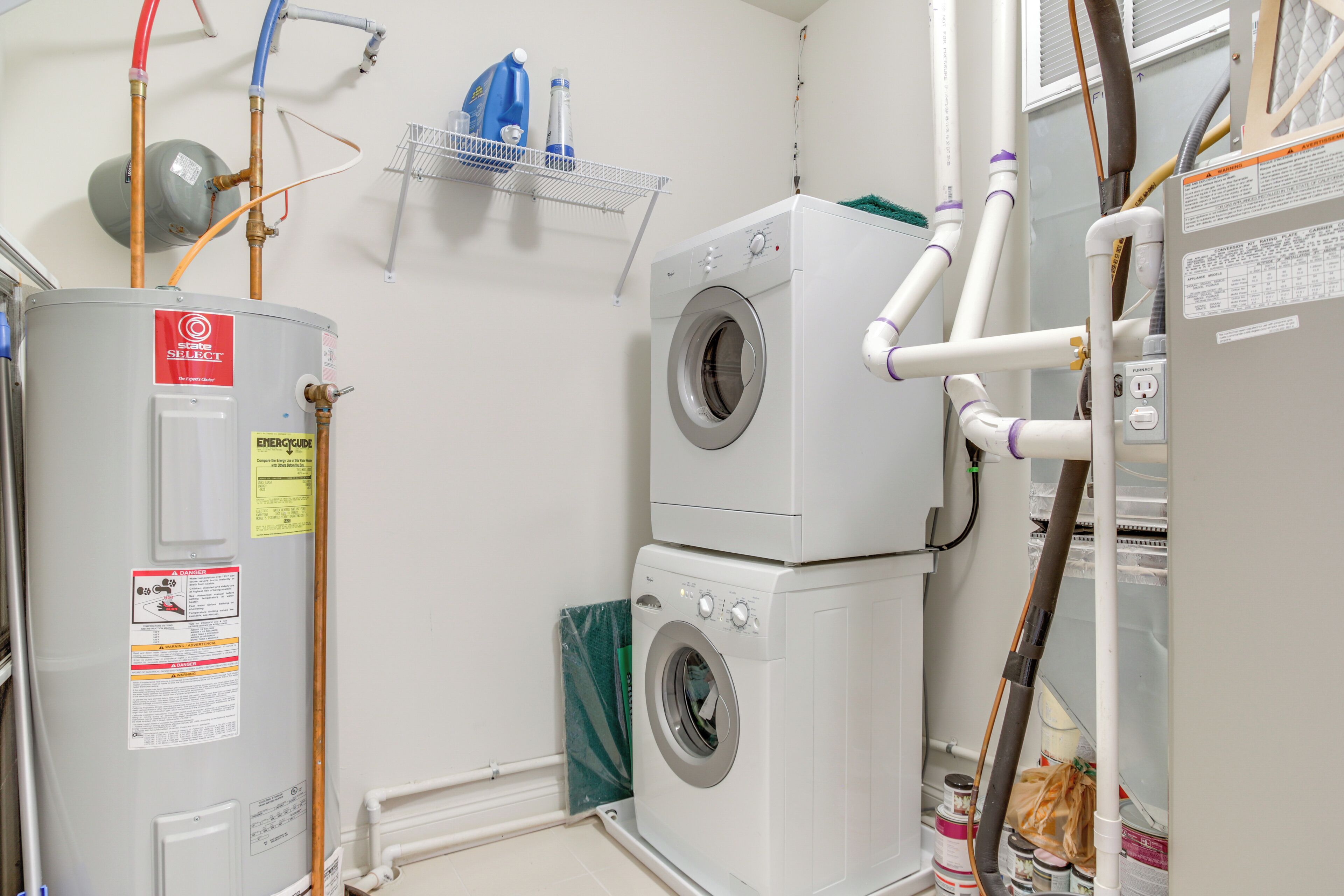 A laundry room with a stacked washer and dryer, a water heater, and various pipes and hoses.