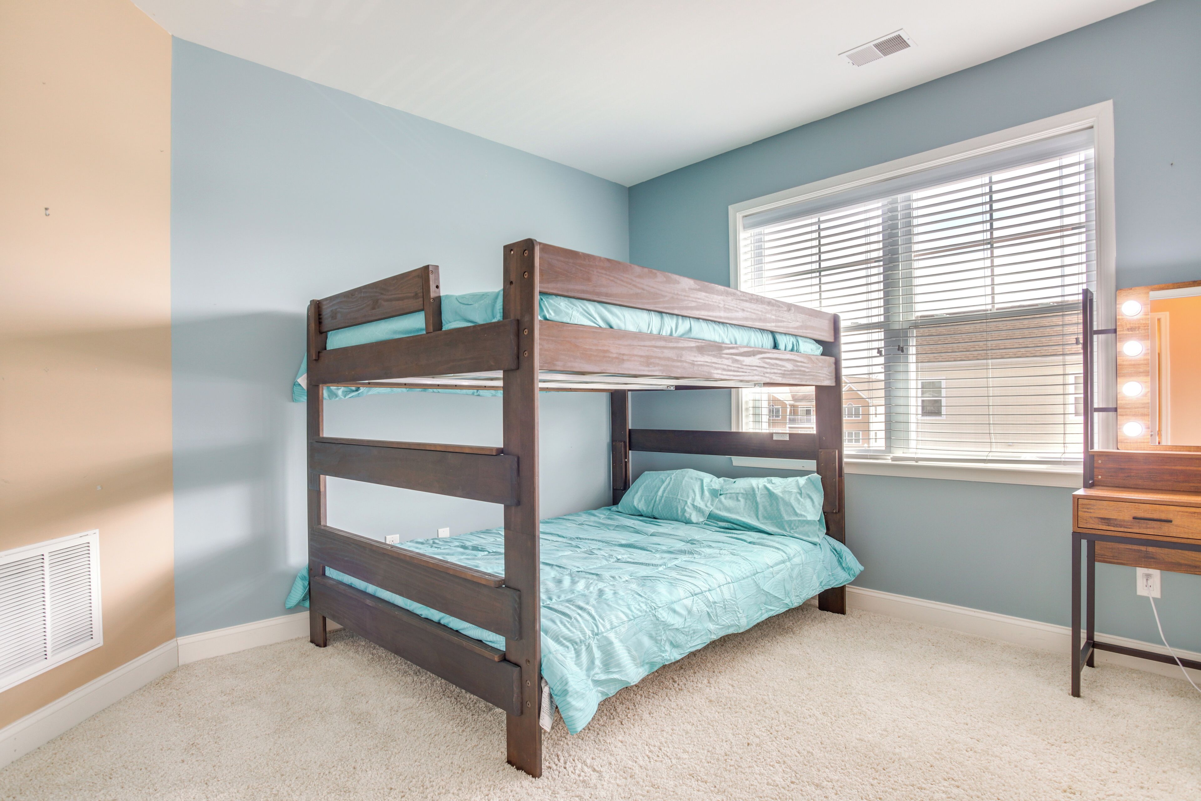 Un lit superposé dans une chambre aux murs bleu clair, avec une fenêtre équipée de stores et un petit bureau agrémenté d’un miroir.