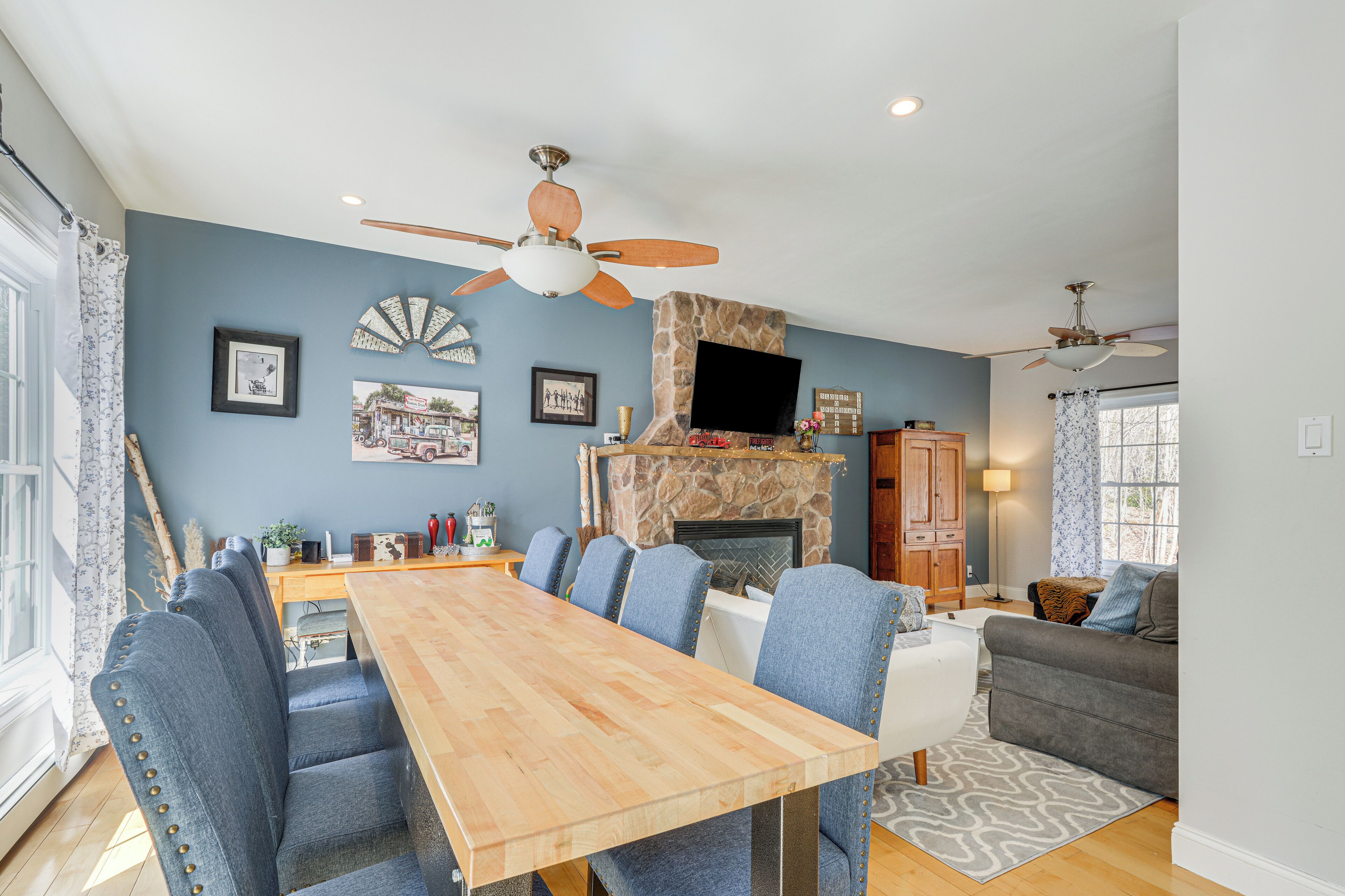 Een ruime woonkamer met een open haard, een eethoek met een houten tafel en blauwe fauteuils, en een grijze bank.