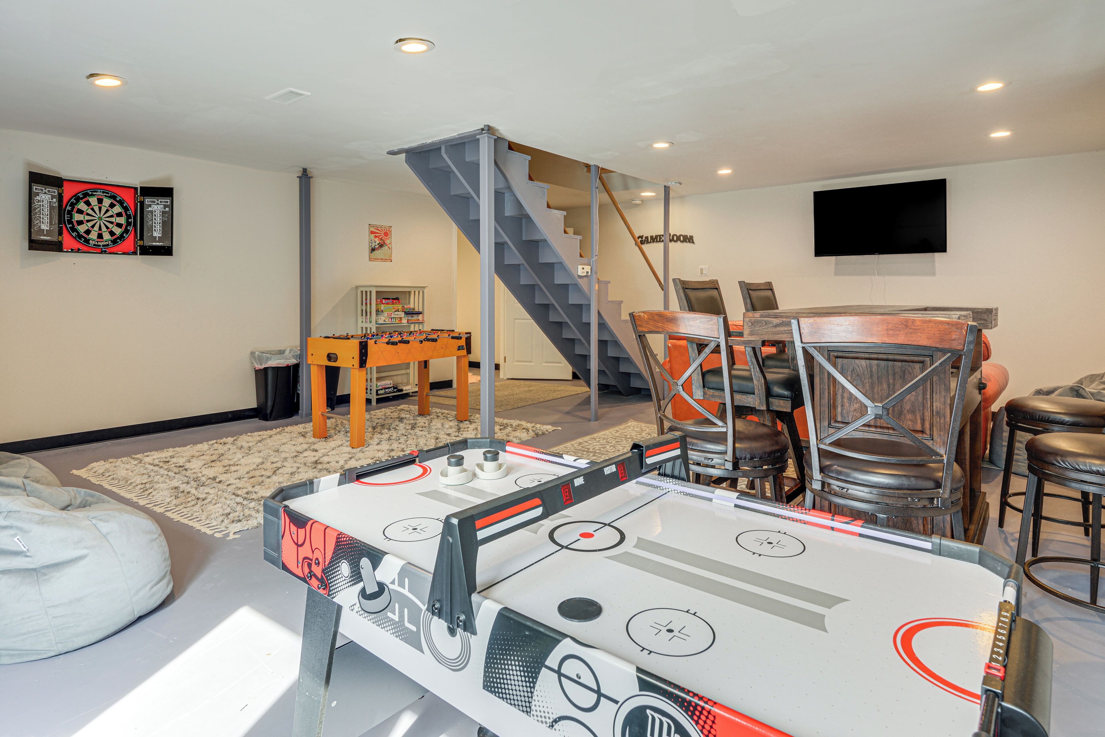 A living room with a foosball table, a dartboard, a TV mounted on the wall, and a staircase.