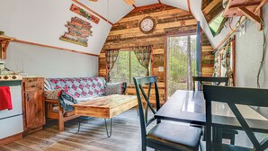 Un interior de cabaña acogedor con techo de madera, una mesa de comedor con sillas, un sofá con cojines estampados y una cocina con estufa y refrigerador.