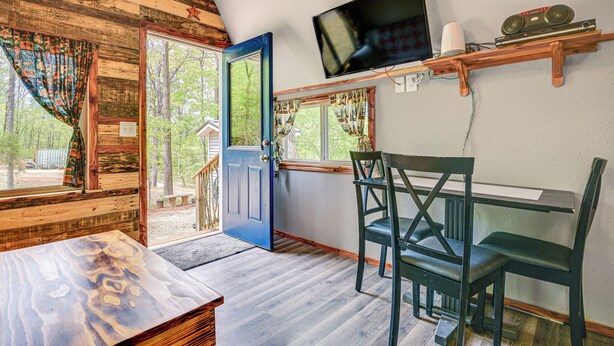 Interior rústico de una cabaña con techo de madera, una puerta azul, un televisor montado en un estante de madera y una mesa de comedor con sillas.