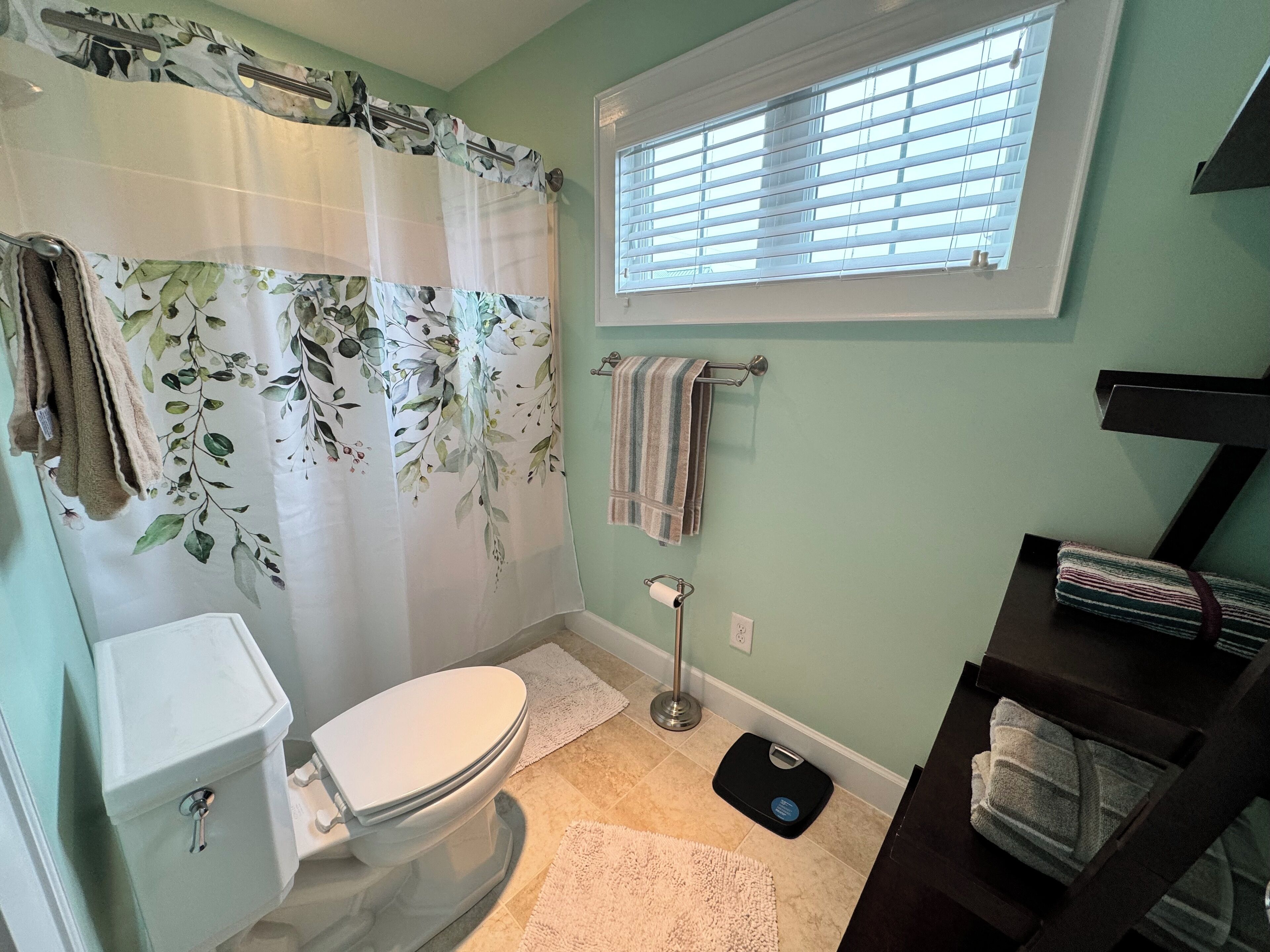 Un baño con inodoro, cortina de ducha estampada con motivos florales, ventana con persianas y toallero con toallas.