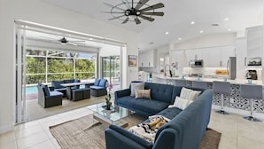 A modern living room with a blue sofa, a glass coffee table, and a dining area with a kitchen in the background. - 5BR Marco Island Home | Pool & Hot Tub (Marco Island)