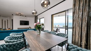 A modern dining area with a wooden table and tufted chairs, a large window with a view, and a television on the wall. - Luxury Villa Brajdini (Vrbnik)