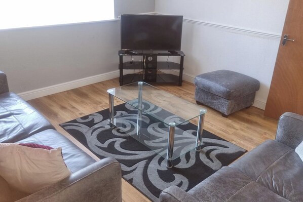 A living room with a glass coffee table, grey sofas, a grey ottoman, a flat-screen TV, and a patterned rug. - Doncaster Dream House Retreat- Sleeps 9 (Doncaster)