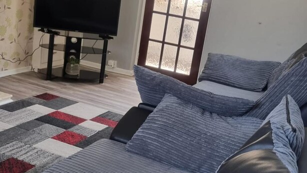 A living room with a sectional sofa, a flat-screen TV on a stand, a patterned rug, and a window with frosted glass. - Doncaster-house-sleeps8-parking-garden (Doncaster)