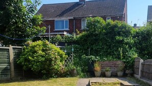 A residential backyard with a paved pathway leading to a wooden deck, surrounded by lush greenery and a brick house in the background. - Doncaster-house-sleeps8-parking-garden (Doncaster)