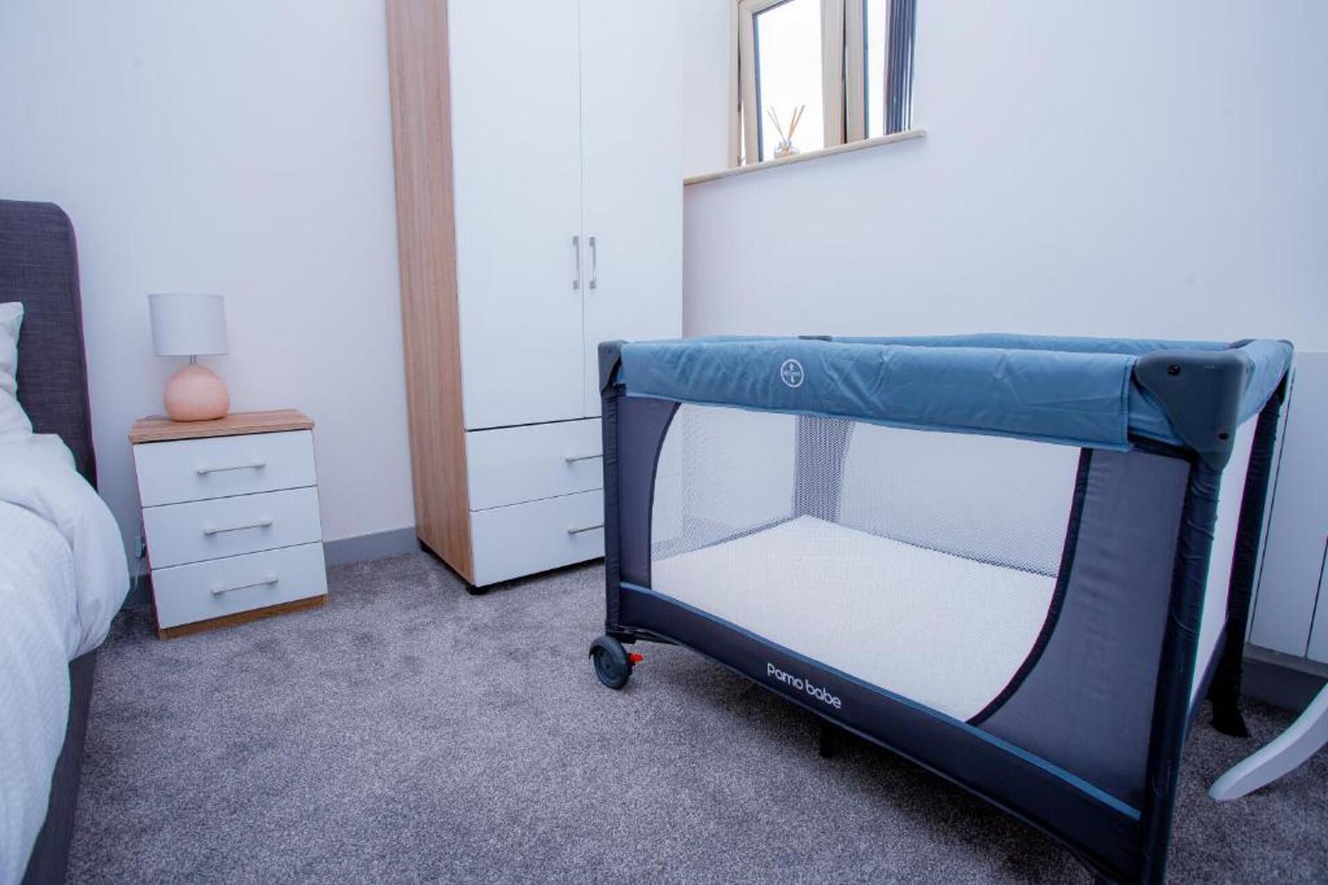 A baby playpen in a bedroom with a bed, dresser, and a window.