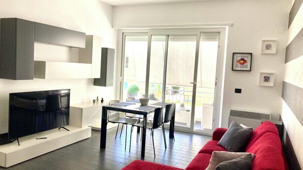 A modern living room with a red sofa, a flat-screen TV, a dining table, and a sliding glass door leading to a balcony. - Casa vacanza a due passi dal mare, centralissima, ristrutturata (Terracina)