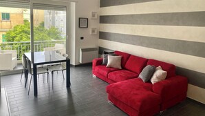 A modern living room with a red sofa, a dining table with chairs, and a large window overlooking a balcony. - Casa vacanza a due passi dal mare, centralissima, ristrutturata (Terracina)