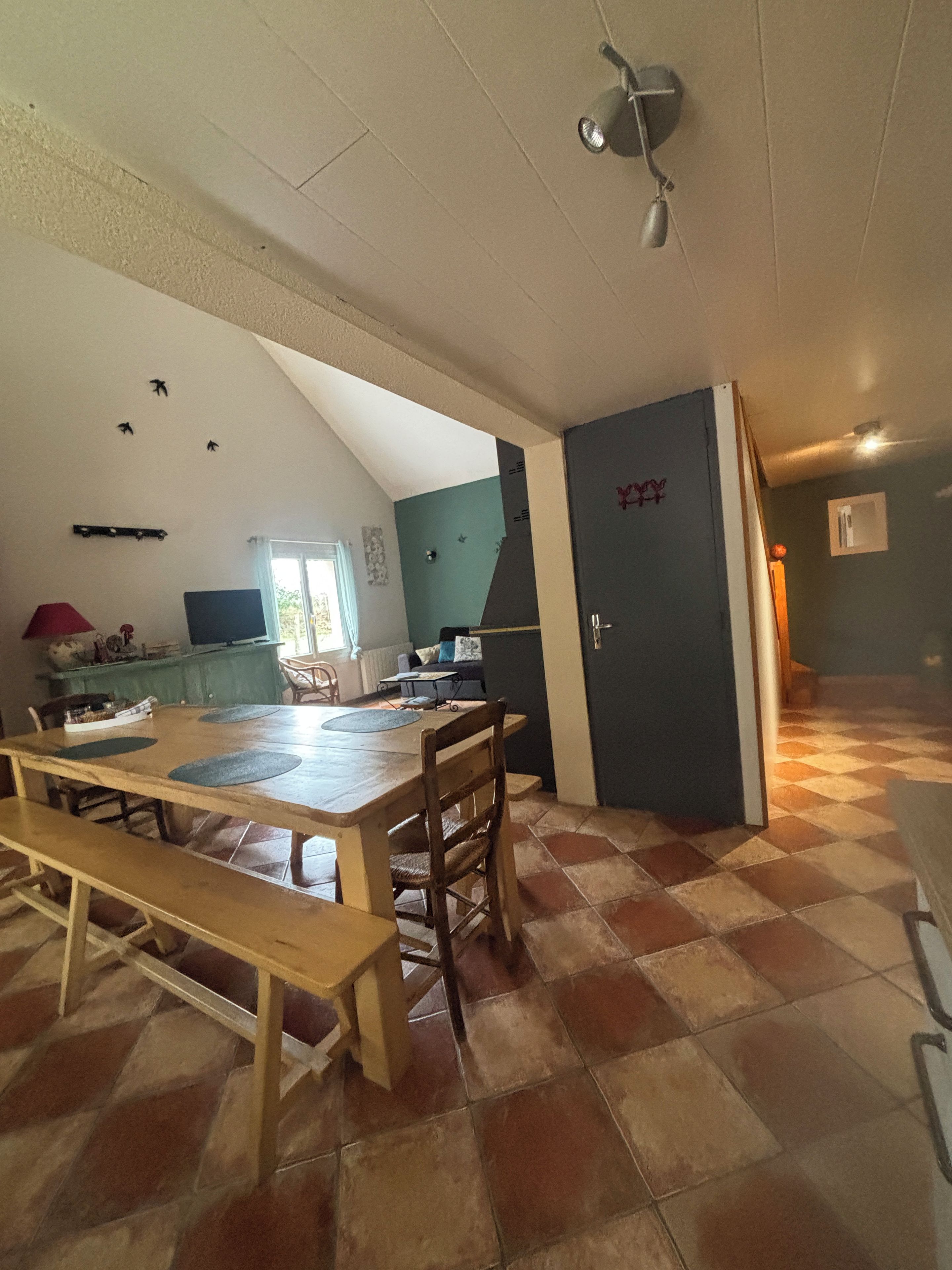 A dining area with a wooden table and benches, a kitchen in the background, and a tiled floor.