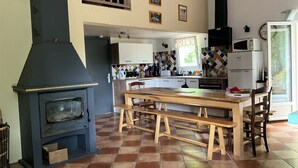 A kitchen with a wood-burning stove, a dining table with benches, and a window with a view of greenery.