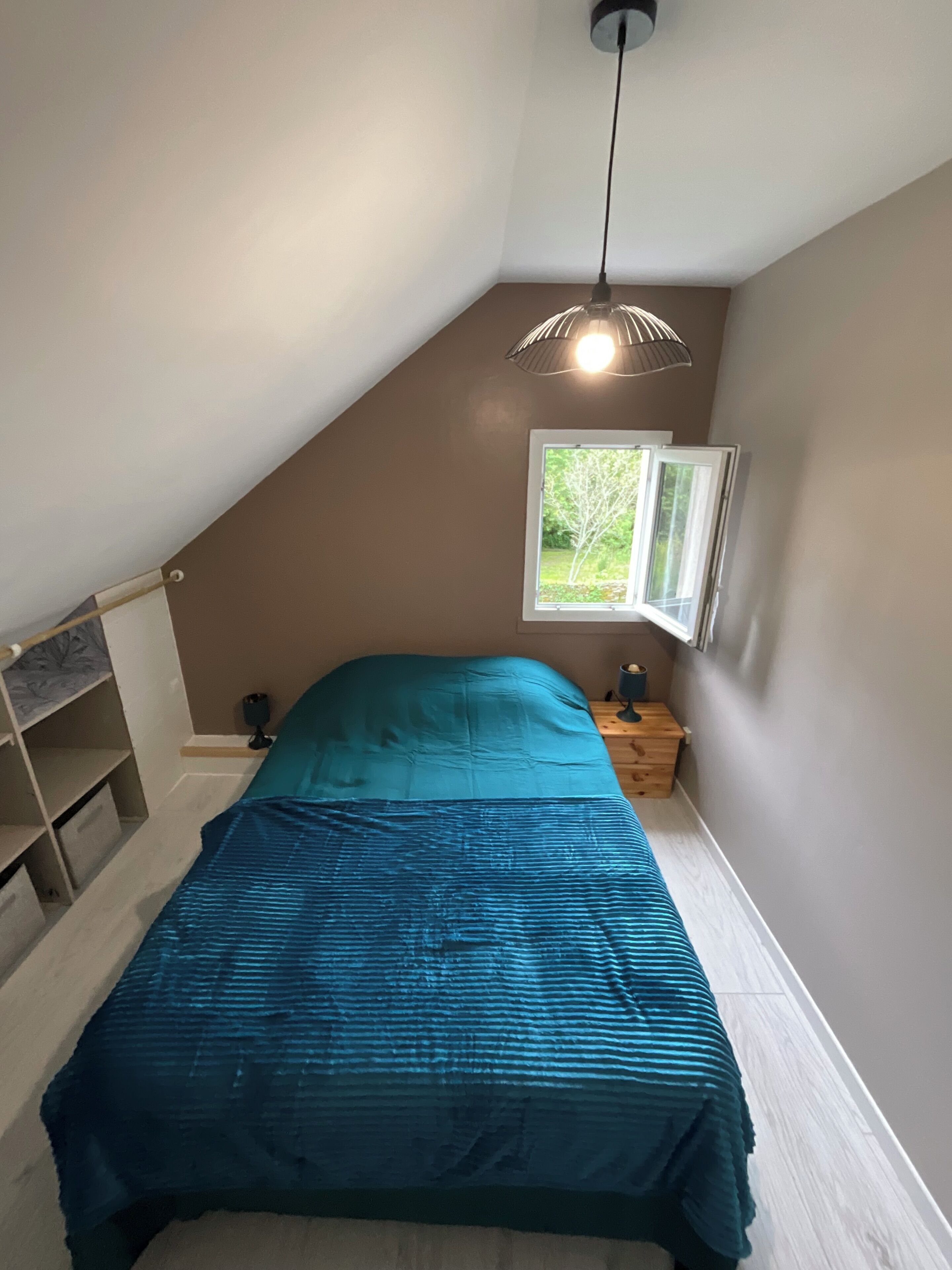 Ein Schlafzimmer mit schrägen Wänden, einem Bett mit türkisfarbenem Bettbezug, einem Fenster mit Blick auf Grünflächen und einem Nachttisch mit Lampe.