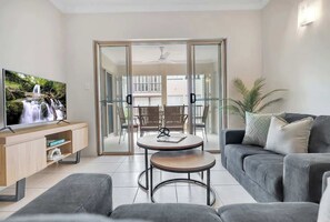Een moderne woonkamer met een flatscreen-tv, een grijze bank, een klein rond tafeltje en een glazen deur die leidt naar een buitenruimte met een stoel en een plant.