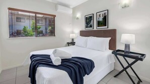 A hotel room with a bed, two bedside lamps, a ceiling fan, a window with blinds, and two framed pictures on the wall.