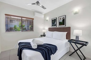 A hotel room with a bed, two bedside lamps, a ceiling fan, a window with blinds, and two framed pictures on the wall.