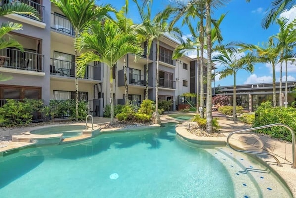 A hotel with a swimming pool surrounded by palm trees and a multi-story building with balconies.