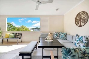 Une pièce avec vue sur la verdure, un ventilateur de plafond, une table avec un vase, un canapé avec des coussins à motifs et un banc.