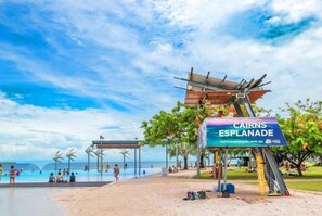 A beach with a sign reading 'CAIRNS ESPLANADE,' a pool area, and a pavilion structure.