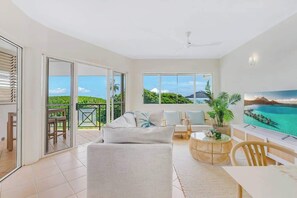 A spacious living room with a large sliding glass door leading to a balcony, a comfortable sofa, a dining table, and a television.