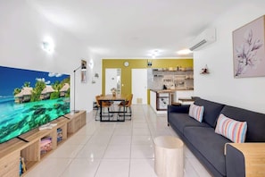 A modern living room with a flat-screen TV, a sofa, a dining table, and a kitchen in the background.